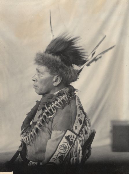 Profile studio portrait of Gegwejiwebinung or Gaygwachewaybinanung (Trying To Throw) or Gayshiqonnayyash (Swift Feather), Called Red Blanket, with Headdress and Bear Claw Necklace. Part of Algonquian and Chippewa (Leech Lake) Tribes.