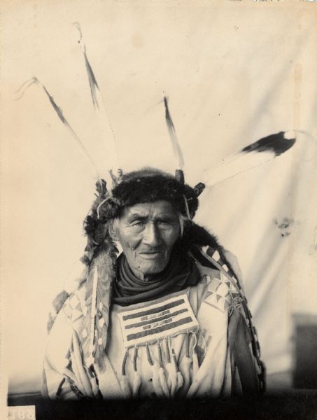 Portrait (Front) of Gemiwunac or Gaymewahnash (Bird That Flies Through the Rain) with Headdress and Holding Pipe-tomahawk. Part of Algonquian and Chippewa (Leech Lake) Tribes.