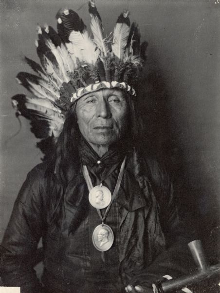 Portrait of Yankton Dakota man, Hinhancaga Maza, (Iron Owl). Part of Siouan (Sioux) and Yankton Tribes. He wears two medals and holds a pipe.