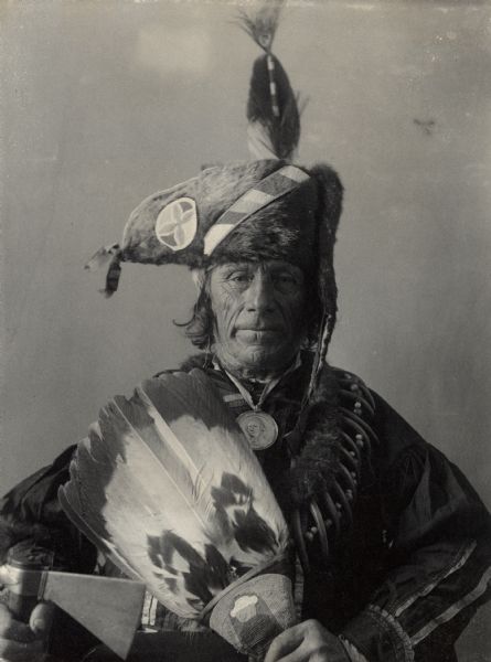 Studio portrait of Oto man, Mato Nayin or Standing Bear, wearing a medal, and holding an axe and fan. Part of Siouan (Sioux) and Otoe Tribes.
