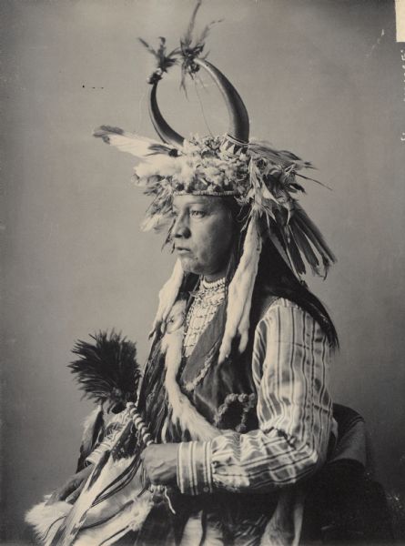 Studio portrait (Profile) of Chief Paul Showeway with horned headdress and ornaments and holding a fan. Part of Waulatpuan and Cayuse Tribes.