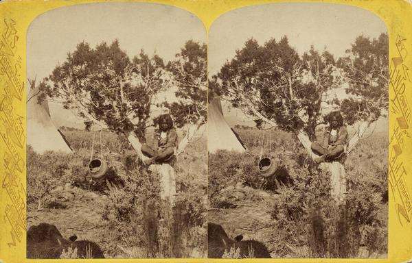Stereograph of a Native American boy, a Ute Indian, seated in a small tree. There is a piece of pottery hanging from a branch of the tree, and a tepee is in the background.