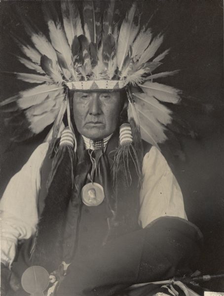 Studio portrait of Shon-Ge-Hin-Ci (Yellow Horse), Brother of Mon-Chu-Non-Zhin (Standing Bear) in partial native dress with peace medal and headdress. Part of Siouan (Sioux) and Ponka Tribes.