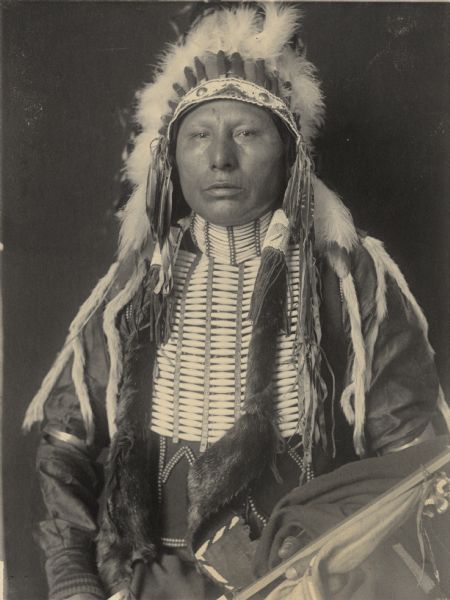 Studio portrait of Shon-Ge-Ni-Ka-Ga-Hi (Horse Chief), Son of White Eagle, in partial native dress with headdress, breastplate and holding pipe. Part of Siouan (Sioux) and Ponka Tribes.