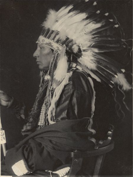 Profile studio portrait of Shon-Ge-Ni-Ka-Ga-Hi (Horse Chief), Son of White Eagle, in partial native dress with headdress, breastplate and holding pipe. Part of Siouan (Sioux) and Ponka Tribes.