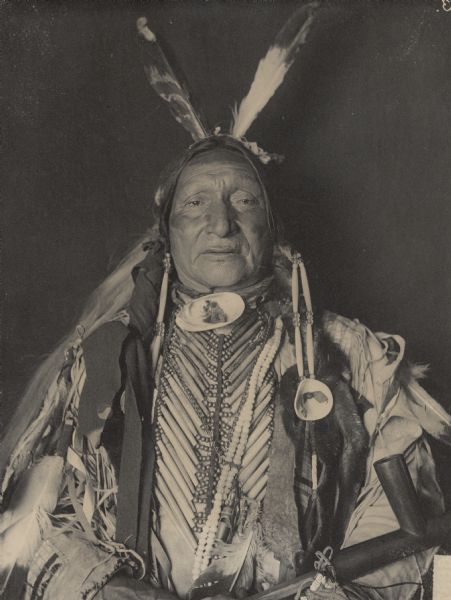 Studio portrait (Front) of Siyo Sapa (Black Chicken) or (Prairie Hen) in native dress with breastplate and holding pipe. Part of Siouan (Sioux) and Yankton Tribes.
