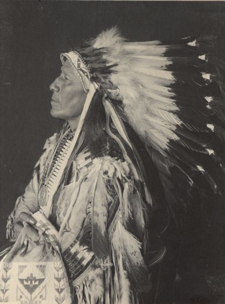 Studio portrait of Chief of Loafer Band, Tasemke-Tokeco, called Paul Strange Horse, in native dress with peace medal, headdress, breastplate and holding bag. Part of Siouan (Sioux) and Brule Tribe.