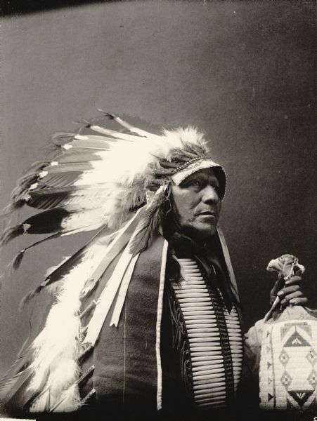 Studio portrait of Chief of Loafer Band, Ta-Semke-Tokeco, called Paul Strange Horse, in native dress with headdress, breastplate and holding bag. Part of Siouan (Sioux) and Brule Tribes.