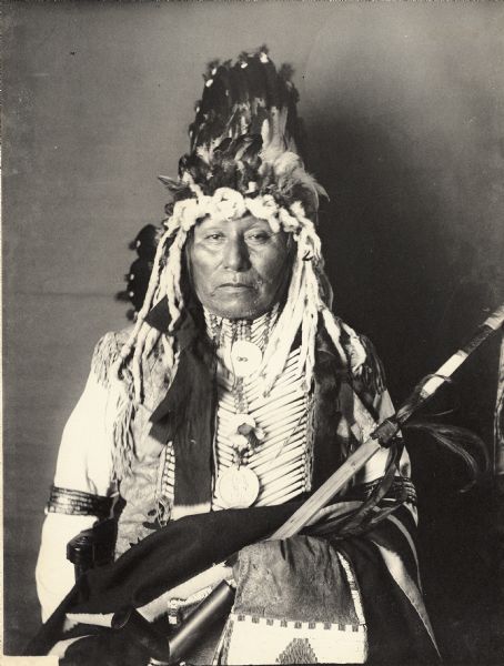 Studio portrait of Wakinyanzi or Wakinyan Pazi (Yellow Thunder) in partial native dress with peace medal, headdress and breastplate and holding a pipe and bag. Part of Siouan (Sioux) and Yankton Tribes.