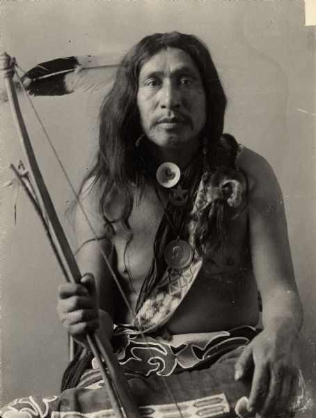 Studio portrait of Wogewashonewahe (Gray Stone), called Robert McGlashen, in native dress with peace medal and holding bow and arrow. Part of Siouan (Sioux) and Otoe Tribes.
