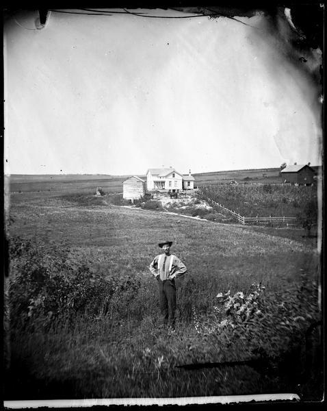 View of the Quammen family farm with a man in the foreground standing in a field. Behind him and across the field is an upright and wing frame house with what appears to be a small summer kitchen. There are two people standing near a log building on the left, which may be the original dwelling converted to agrarian use (possibly a granary) when the frame house was constructed. Another person is standing on the porch of the farmhouse, and two people are on a horse-drawn buggy. A manure pile is between the house and a barn.