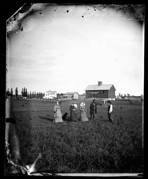 H. Sveren farm with the family standing in the foreground and a dog, farm buildings and fences in the background. A line of trees is behind the farmhouse on the left. Walworth Co.