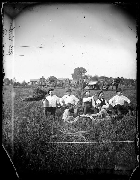 Two girls are sitting and facing each other in a field in front of several men and a boy wearing hats, and a woman standing and holding a fan. A horse-drawn reaper, wagons are being the group, and a farmstead is in the far background.