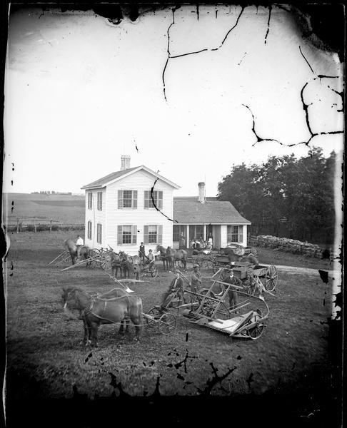 Elevated view of group of people and farmhouse. There are three men with a horse-drawn self-rake reaper, and behind them are more people, several horses and horse-drawn vehicles. An upright and wing house with prominent lightning rods is in the middle ground, with several people on the porch.