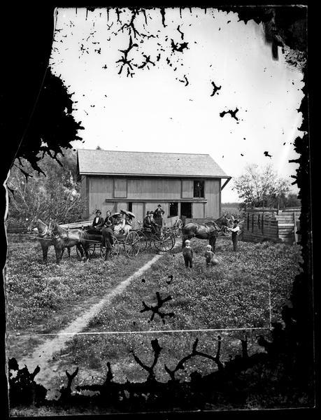 View down farmyard towards a couple sitting in a carriage on the left, and a family, including three girls with parasols, sitting in another vehicle in the barnyard. Two men and children are standing nearby. In the background, a man is standing in a wooden wagon in front of a barn.