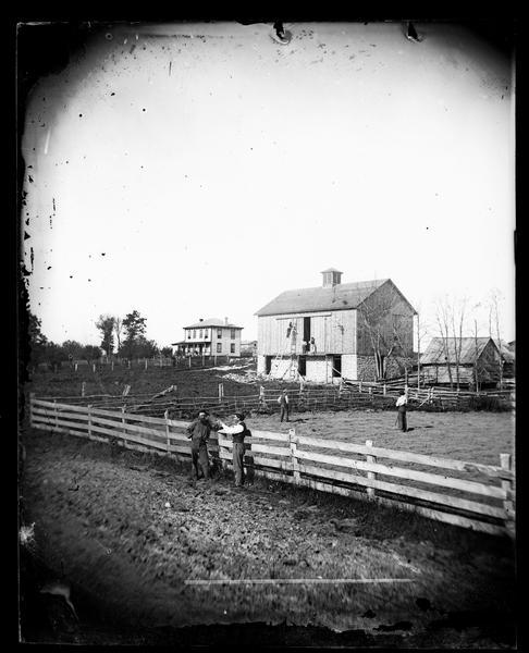 Two men are leaning on a fence in the foreground, with other men behind them in a field. Men on scaffolding and ladders are building a barn with a cupola. Other buildings include a log building on the side of the barn and an Italianate house in the background.