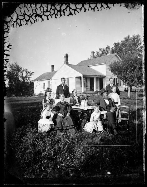 A family in their best clothes sitting around a table in front of their frame house.
