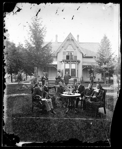 A family is sitting around a table in the yard. Other family members in hats are in the background, standing in front of a frame house with a bay window and gable and cut work at the top.