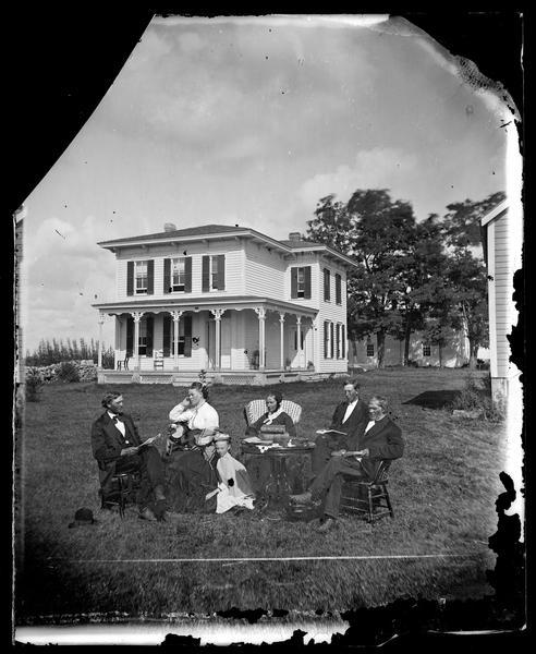 A family posing around a table covered with books and a simple cloth in front of a two-story bracket-style frame house with a wrap-around porch.