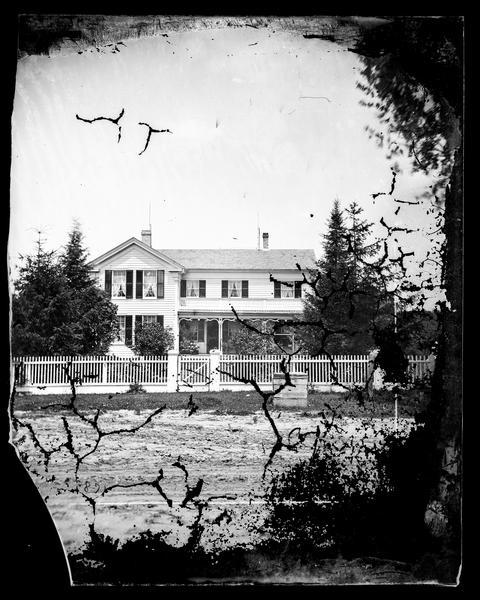 View from the road of a frame house behind a picket fence, with cut work, shutters, and  lightning rods by each chimney and a well in front of the fence. View is possibly of Washington Road in Christiana or 324 South Fifth Street in Stoughton.