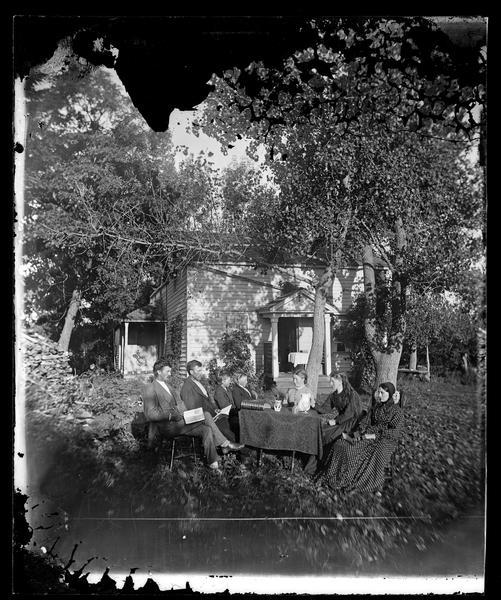 The Newhouse family sitting around a table under trees with a book, goblets, a vase of flowers, a pitcher, a stereoscope and a cloth on the table in front of a frame house. Over the entrance to the house is a sign that reads: "Hekla" indicating that the house is insured by the Hekla Fire Insurance Company. From left to right, they are: Christian Newhouse, Ole Newhouse, unknown, Knut Newhouse, Rachel Larson, Anna Newhouse, and Kari Newhouse.