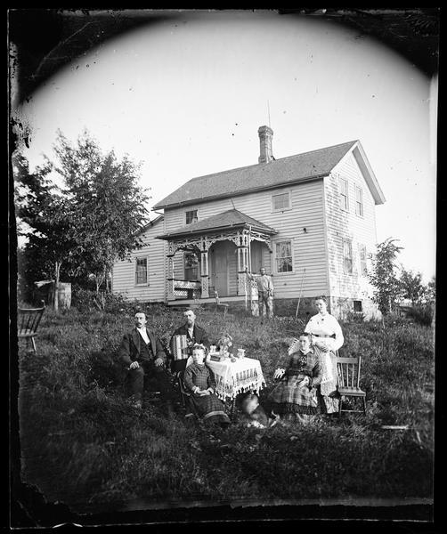 A man with an accordion, another man with a clay pipe, two women and two children are sitting around a table that has a knit cloth, a vase, a book, plants and fruit on it. Another man is standing in the background by the porch of a two-story frame house. The house has carpenters lace on the front porch, cut work above the windows, a stone foundation, and a lightning rod near the chimney.