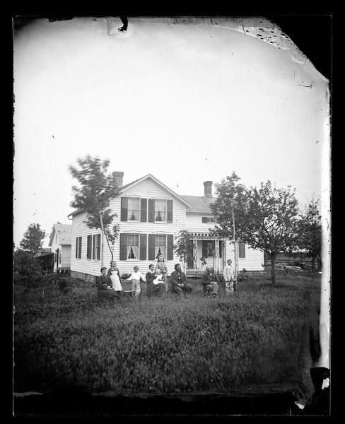 A family sitting in a row in front of their upright wing frame house. The house has cut work on the porch, small windows above the porch and a stone foundation.