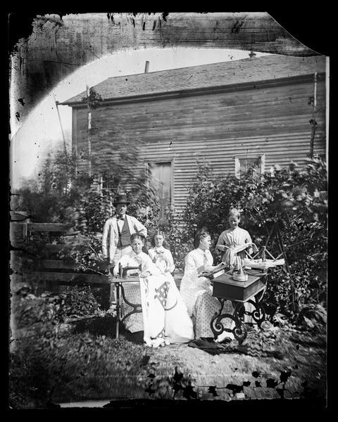 The G. Thompson family posing in front of their wooden house.  One woman is sitting at a sewing machine working on a quilt while another is working at a knitting machine.