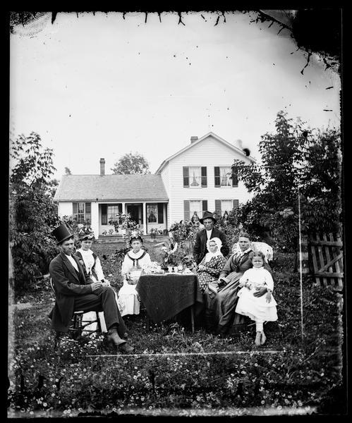 A family is sitting around a table with a cloth, vases and other objects on it. The man on the left is wearing a top hat, and a flower in his lapel. The two women next to him are wearing decorated hats. The woman on the right is holding a stereoscope. Flowers are growing in the yard where the group is sitting. The family's upright and wing frame house is in the background.