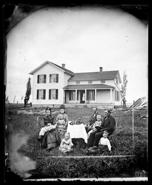 A family is sitting around a table with a stereoscope on it. An upright and wing frame house with a stone foundation is behind them. The woman has a large doll laid across her lap.