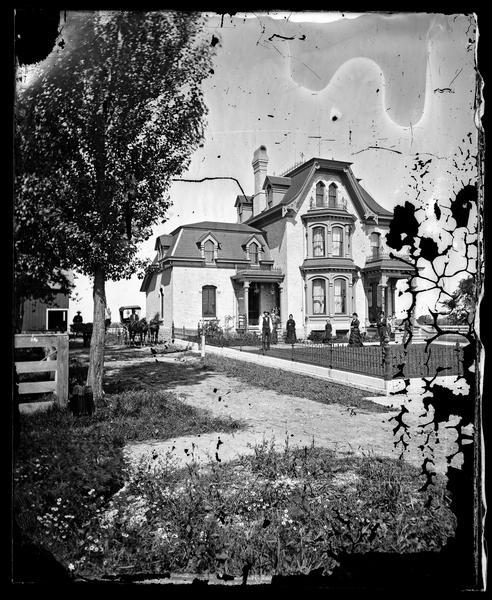 The Martin K. Dahl family is in the yard of their brick Victorian home, which is surrounded by an iron fence. The house has iron work on porch tops and on the roof.