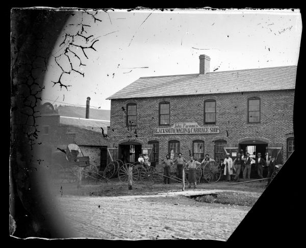 John Parman's Blacksmith, Wagon & Carriage Shop. A large group of men, including workers in leather aprons, are standing with carriages and wagon wheels outside the shop. Three children are sitting in a wagon on the left.