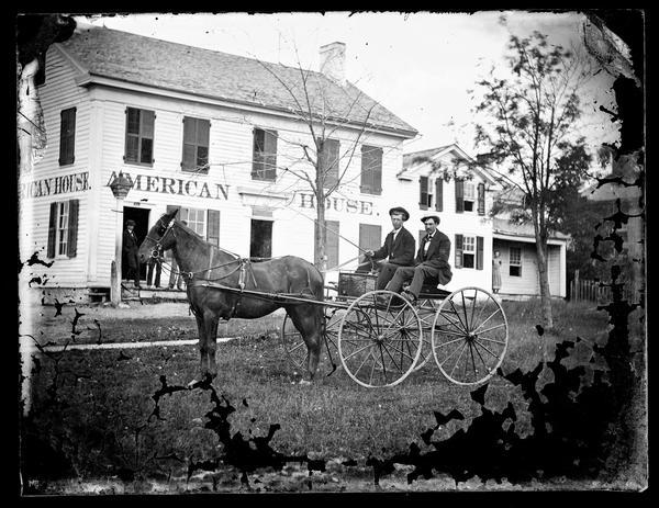 Two men are sitting in a carriage in front of a Madison hotel known as both the Flom Hotel and the American House. The Flom Hotel, built in 1865, was located on the corner of South Hancock and East Main Streets. It should not be confused with the American House hotel located on the Capitol Square on Pinckney Street at the corner of Washington Avenue, which was built in 1838 and burned in 1868. The hotel was owned by the Jefferson family, and the men in the buggy have been identified as Beverly Jefferson, sitting on the left and his brother, John Jefferson, on the right.