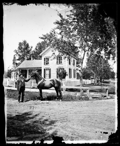View of Dr. Blake's (probably Samuel M.) house in Lodi or Prairie du Sac with a white picket fence in front of it. A man is standing in the grass in front of the sidewalk with a horse, Reaper Boy, identified by a small sign on the road in the foreground. Another man and a woman are standing near the house in the background.