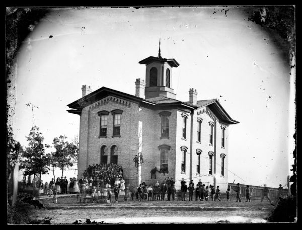 View of a brick school building and a crowd of students, with two boys up in a tree. Other people are posing in some of the open windows of the school. Boys on the right appear to be playing rough games with sticks.