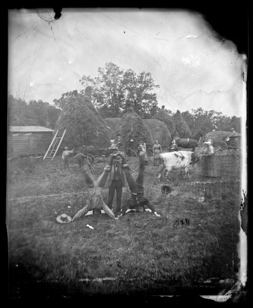 In the foreground, two men are doing headstands while another man is holding their legs together over his head. Women and children are watching from behind, near two horses and a cow. In the background are large, tall haystacks.