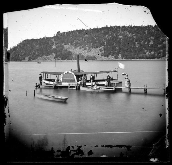 Elevated view of people, elegantly dressed, posing on a pier, with a steamboat and rowboats docked on it on Devil's Lake. An American flag is on the steamboat. In the background is a bluff along the shoreline.