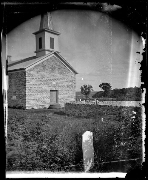 View of the East Koshkonong Church, a stone church built in 1858 and rebuilt in 1893. One grave marking and a stone fence are in the foreground. A brick wall and a gate are near the front entrance.