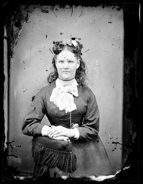 Three-quarter length portrait of a young woman with bows in her hair, a choker locket, a fringed scarf around her neck and a pocket watch around her waist. She is leaning on an armpost with tassels.