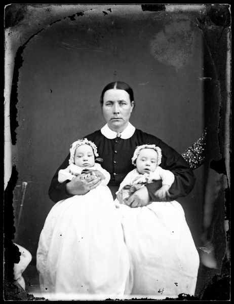 Portrait of a woman holindg two babies in her lap. The babies appear to be twins, and are wearing caps and gowns.