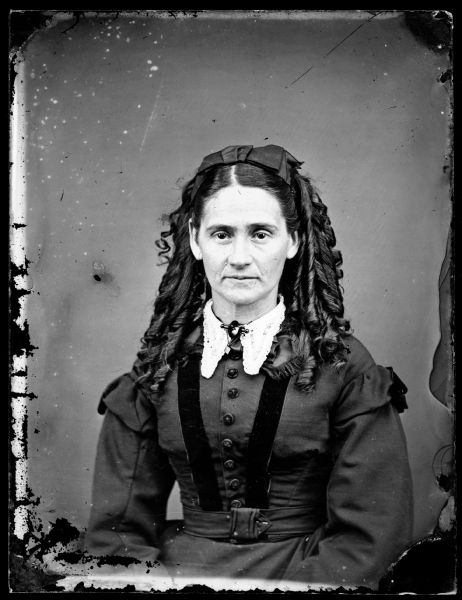 Waist-up portrait of a woman with a lace collar and pin. The woman's hair is in ringlets, and she is wearing a bow on top of her head.