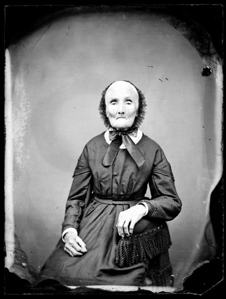 Three-quarter length portrait of an older woman wearing a bonnet sitting in a chair with a tasseled armrest.