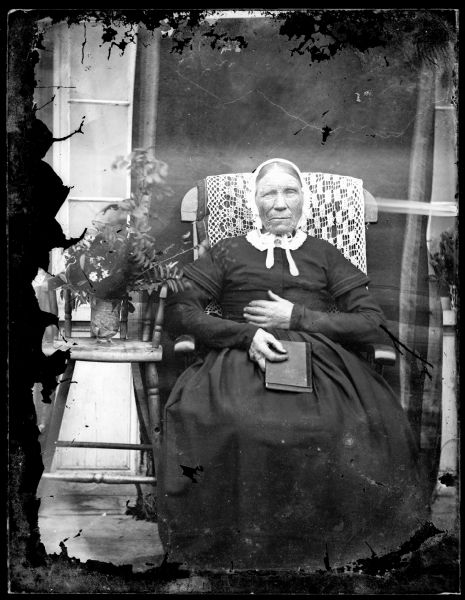 Full-length portrait of an older woman sitting in a chair with a lace cloth on the back. The women is wearing a bonnet and is holding a book in her lap. A vase of flowers is next to her on a stool.