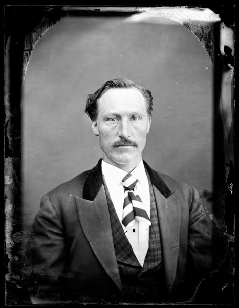 Waist-up portrait of a moustached man in a velvet collared jacket, plaid vest and a striped tie.