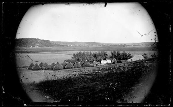View across field towards the Amund Rustebakke farm. Twelve haystacks dominate the left and center of the image, and two people are standing on top of the haystack nearest the house. A group of people are standing next to the frame house, with the village of Black Earth in the distance and bluffs beyond.