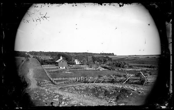 View down a rocky hill towards a split-rail fence bordering a prosperous farm. The low building in the center is the kind of cattle shed settlers built as their first buildings. Circular haystacks in the background are a Northern European feature, and there is a barn with a cupola and a farmhouse. Three people are posing in the barnyard near a horse-drawn wagon on the right, and another man is sitting in a cart on the left side of the barn. A long fence line is along the hill in the background on the right above the farmhouse.
