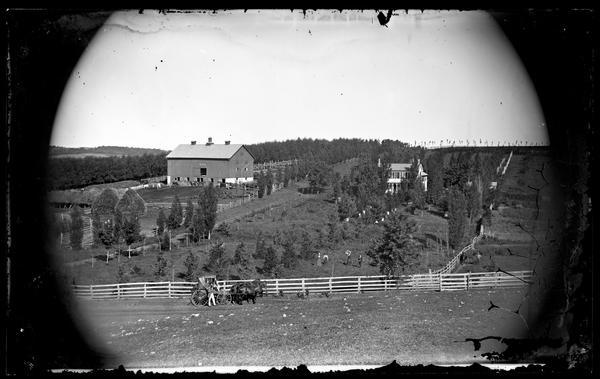 Three people are posing in the foreground, with a man with a long beard standing in front of two women sitting in a horse-drawn carriage. There is a barn and house uphill in the background surrounded by trees. Another group of people, a man and three women, are behind a fence on the right, with three of them holding croquet mallets, and a woman holding an umbrella. Another man is standing on the far right with a horse behind another fence. There are beehives among the shrubs and trees near the farmhouse. A tree-lined lane and extensive fencing runs through the farmstead. Haystacks are on the left near a group of men standing with horses, cows and pigs near the barn.