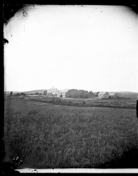 View across field towards farm buildings, with split-rail and other types of fencing surrounding a farmhouse, a barn, a log structure and haystacks. A group of ducks or geese are in the farmyard near the house. Hills are in the background on the left.