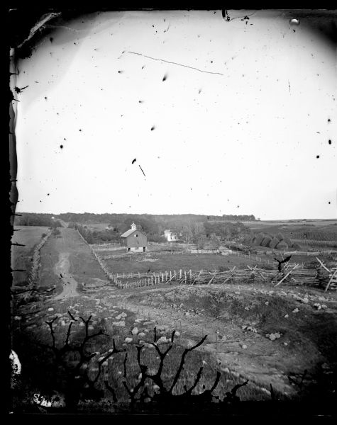 View down a rocky rural lane near West Blue Mounds. Zigzag stake and rider split-rail fencing enclose a farmstead, which includes a barn, a frame house, haystacks, a cornfield, straw-topped shelters and other minimal animal shelters. Two men are standing near a cart on the left side of the barn.