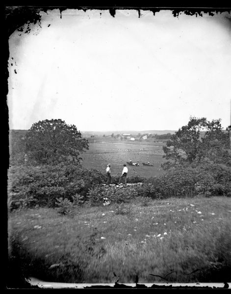 View down hill towards two men in the foreground standing on top of a stone fence looking out onto fields where two or three men are working with two wagons. In the fields are haystacks and a group of animals grazing. The Tollef Gjermundsen farmstead is in the far distance.